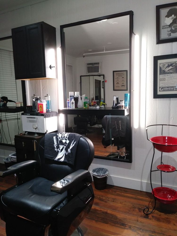 Black barber chair in front of an illuminated mirror, with vintage shaving prints and a red stand